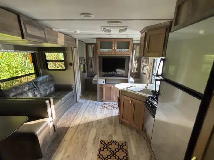 Inside an rv rental, showing the living area with sofa, dinette, and TV. A kitchen with fridge and stove is on the right, with trees visible outside.. Keystone RV Sprinter Campfire Edition 2018