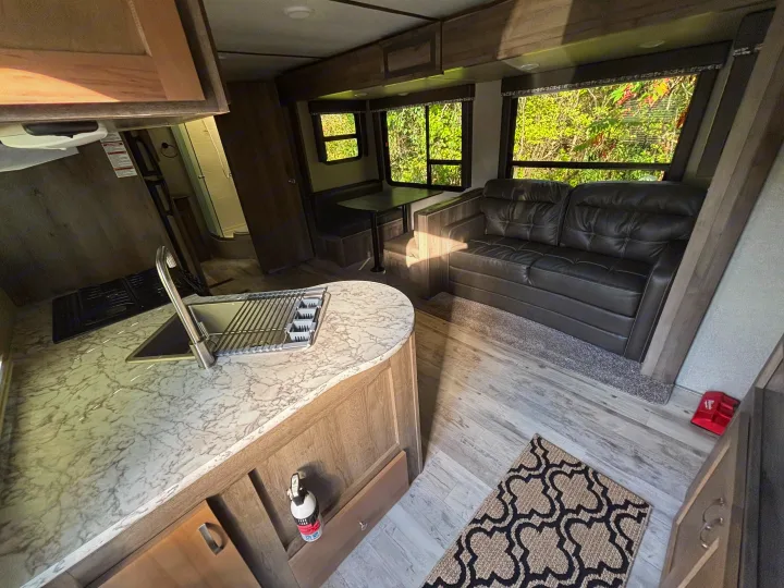 Inside an RV rental: full kitchen with sink, dinette, and cozy living area with a dark sofa. Bright windows show green nature.. Keystone RV Sprinter Campfire Edition 2018