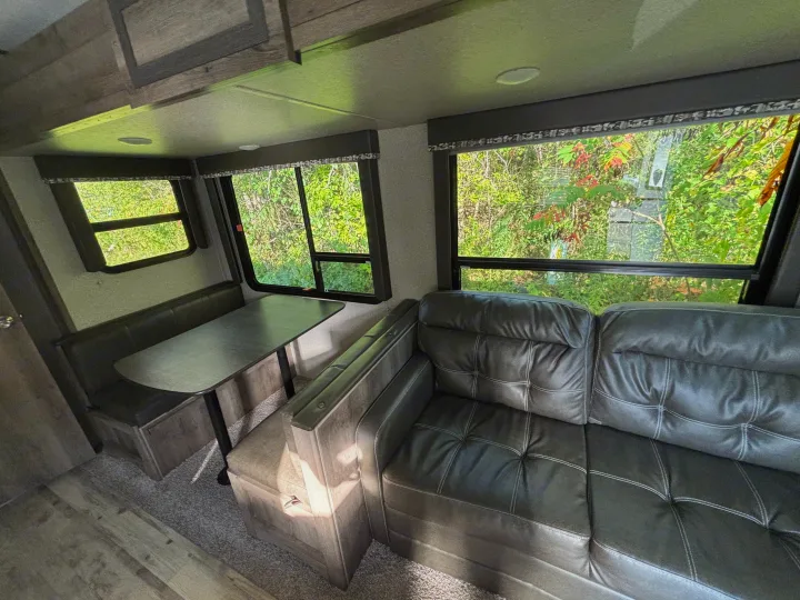 RV rental interior with a cozy dining booth, sofa, and large windows offering lush green views.. Keystone RV Sprinter Campfire Edition 2018