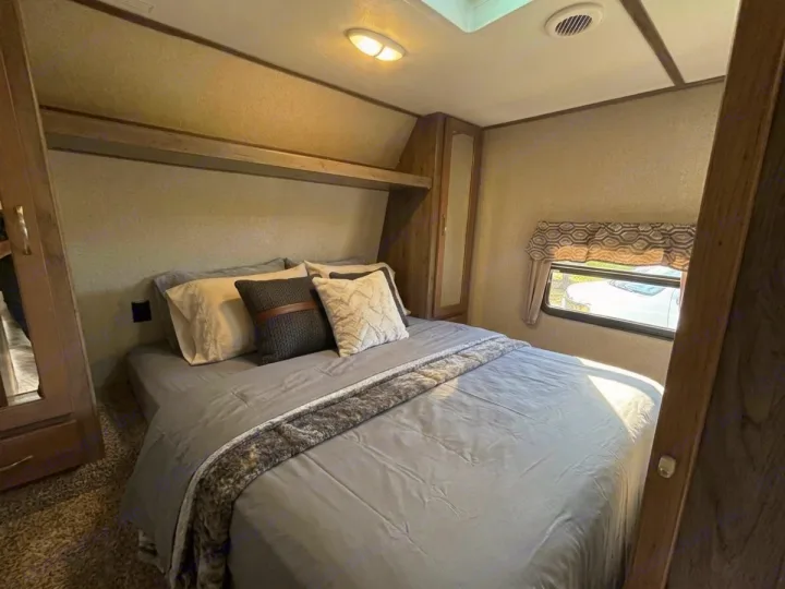 Relax in this comfortable RV rental bedroom. Features a queen bed with gray bedding, pillows, overhead shelf, and a window.. Keystone RV Sprinter Campfire Edition 2018