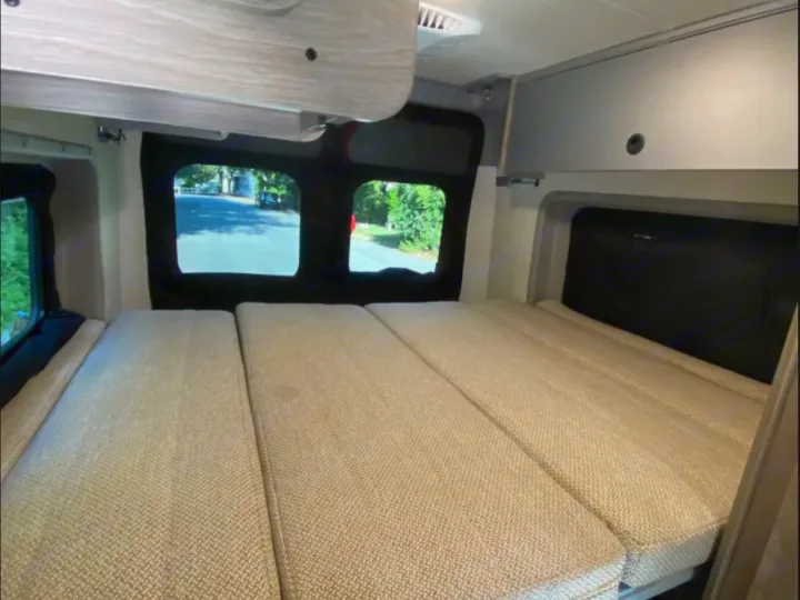 Spacious RV rental interior showing a comfortable modular bed, overhead cabinets, and bright windows offering outdoor views.. Winnebago Solis 2024