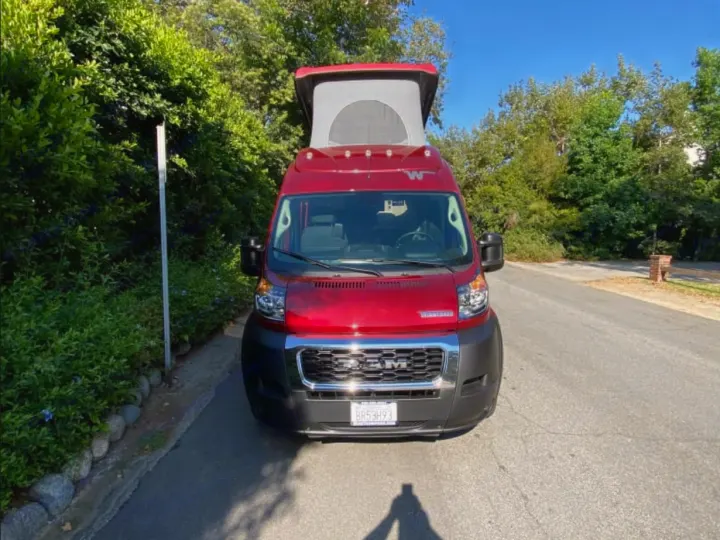 A red Winnebago pop-top camper van, perfect for RV rental, parked on a sunny street with lush greenery.. Winnebago Solis 2024
