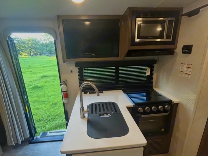 RV rental interior: a clean kitchen with a sink, stove, microwave, and TV, plus an open door revealing a green field.. Rockwood Mini Lite 2104S 2023