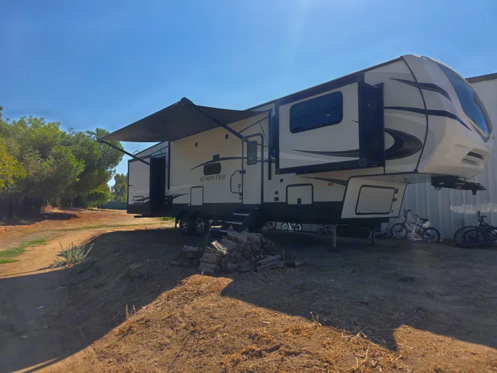 This fifth-wheel rv rental features an extended awning, parked on dirt with firewood & bikes, ready for outdoor trips.. Keystone RV Sprinter Limited 2021