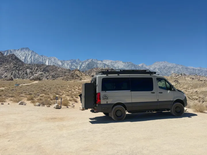 An RV rental van, customized for adventure, parked in a scenic desert landscape with majestic mountains under a clear sky.. Exclusive Outfitters Model B 2026