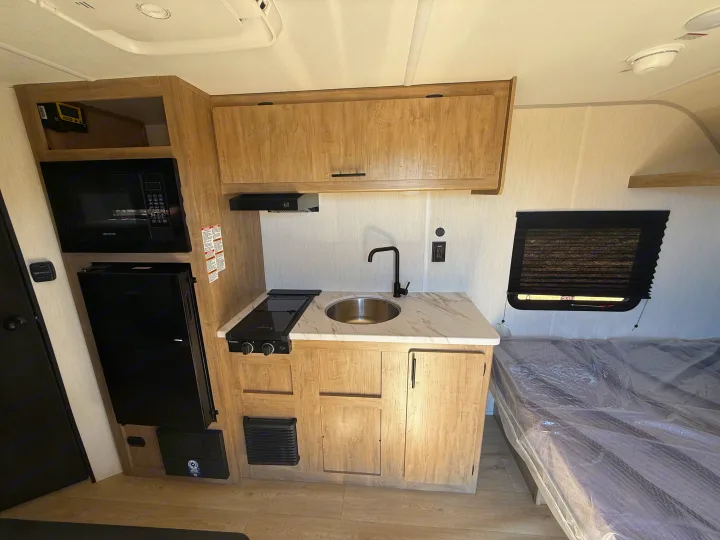Modern RV rental interior with light wood kitchenette: microwave, fridge, stove, sink, and cabinets. A bed is also visible.. Forest River Cherokee Wolf Pup 2026