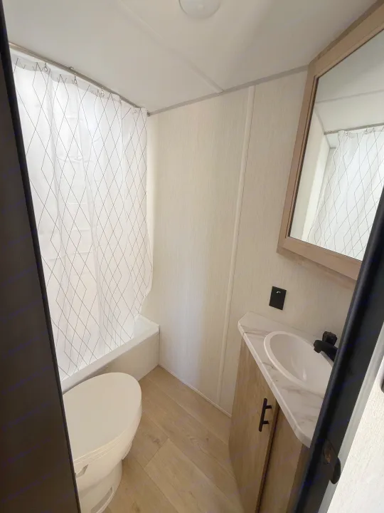 Bright rv rental bathroom with a toilet, shower with patterned curtain, and a modern vanity. Features light wood floors and white walls.. Forest River Cherokee Wolf Pup 2026