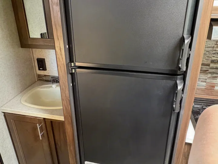 Inside an RV rental: a black two-door refrigerator stands beside a compact bathroom sink with a faucet and vanity.. Forest River Cherokee Grey Wolf 2018