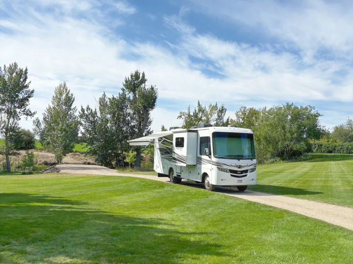 Spacious white RV rental with awning extended, parked on a gravel path amidst lush green lawns and trees under a blue sky.. Jayco Precept 2017