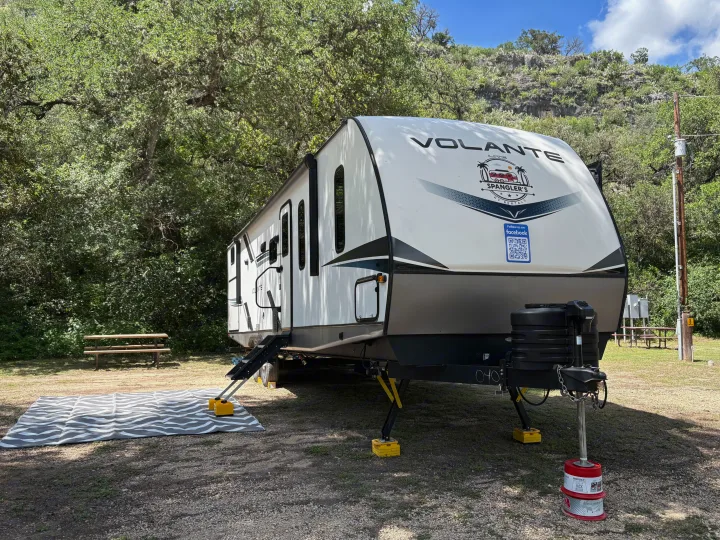 A Spangler's RV rental travel trailer is set up at a scenic campsite with an outdoor mat, picnic tables, and trees.. Crossroads RV Volante 2023