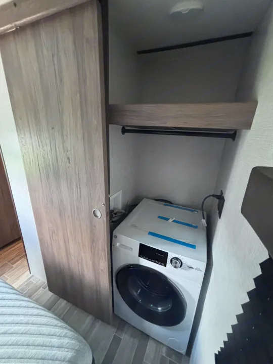 Inside an RV rental: a compact white washing machine is installed in a closet, with a shelf & hanging rod above.. Crossroads RV Zinger 2021