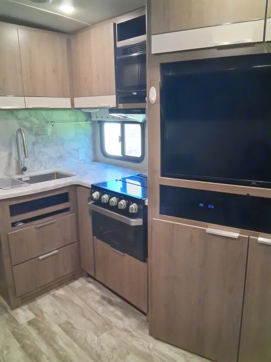 Modern RV rental kitchen with wood-grain cabinets, sink, stove, microwave, and TV showing PM 5:43. Bright & functional.. Grand Design Imagine 2021