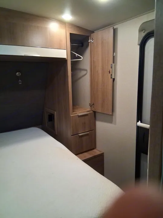 RV rental bedroom view: comfortable bed, open wood wardrobe with hanger, drawers, overhead cabinets, and an entry door.. Grand Design Imagine 2021