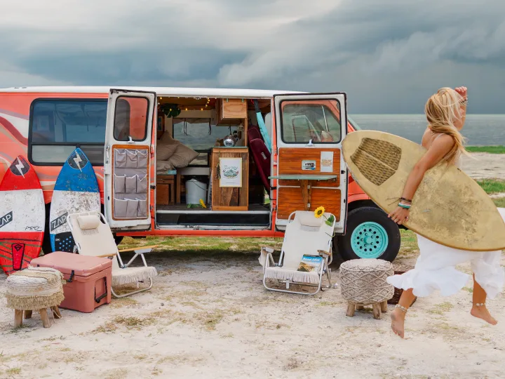 Adventure awaits! A woman with a surfboard jumps by her pink RV rental on a sandy beach, showcasing a cozy van life setup.. Chevrolet G20 Chevy Van 1990