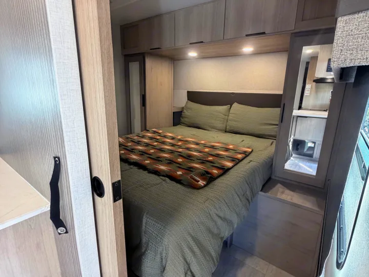 Cozy RV rental bedroom featuring a sage green bed, overhead cabinets with lighting, and a southwestern patterned blanket.. Dutchmen Kodiak 2026