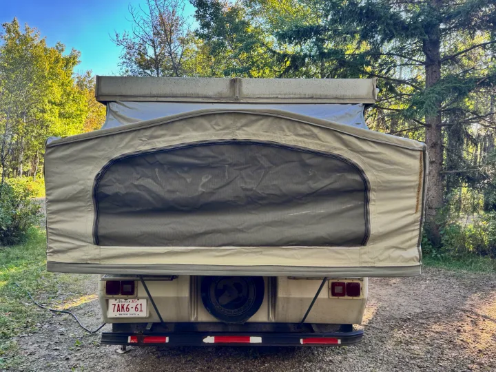 Enjoy nature in this pop-up camper RV rental. Rear view shows large mesh window, license plate, and spare tire in a treed campsite.. Jayco Jay Thrush 6 1989