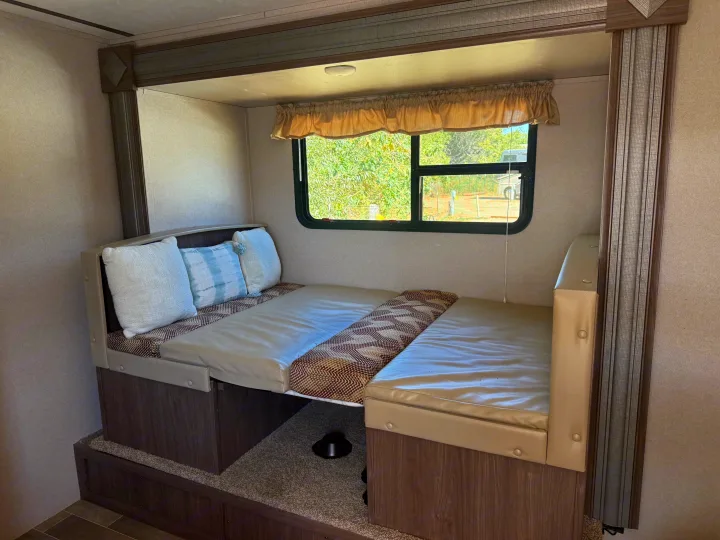 RV rental dinette bed conversion. Cozy sleeping area with pillows, wood trim, and a window view of trees.. Keystone RV Hideout 2020
