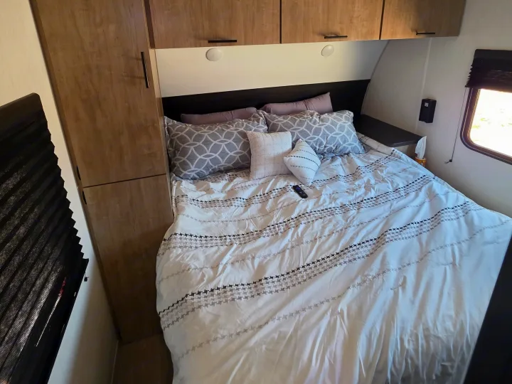 This RV rental offers a compact bedroom with a neatly made bed, white and gray patterned bedding, overhead storage, and bedside amenities.. Forest River Cherokee Grey Wolf 26KF 2026