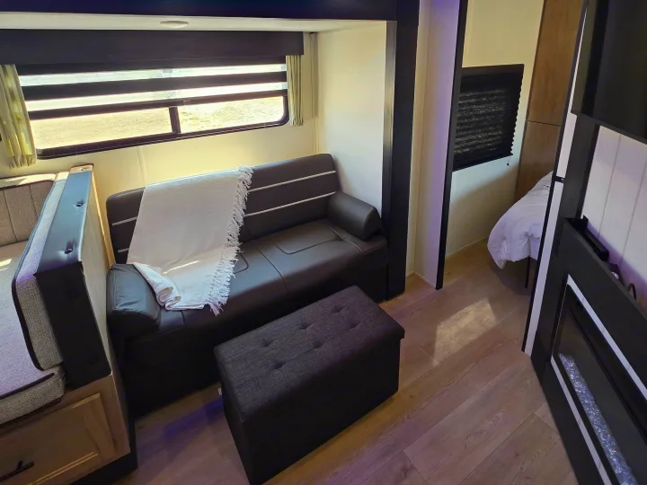 Cozy RV rental living area featuring a dark sofa, ottoman, and a peek into the bedroom. Light wood floors and bright window.. Forest River Cherokee Grey Wolf 26KF 2026