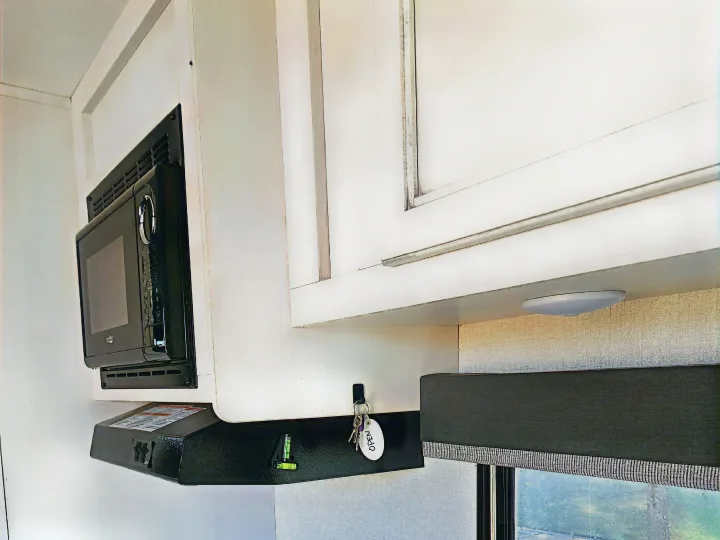 RV rental kitchen: An over-range microwave, vent hood, white cabinets, and a dark window blind are visible. Keys hang on a hook.. Highland Ridge RV Open Range 2021