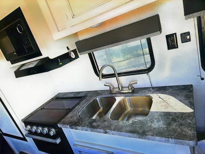 This bright RV rental kitchen showcases a microwave, stove, double sink, and patterned countertop with a window.. Highland Ridge RV Open Range 2021
