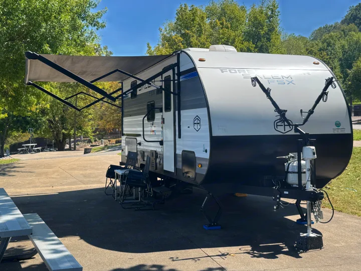 This RV rental is set up at a sunny campsite with its awning extended and outdoor seating arranged on a concrete pad.. Forest River Salem FSX 2025
