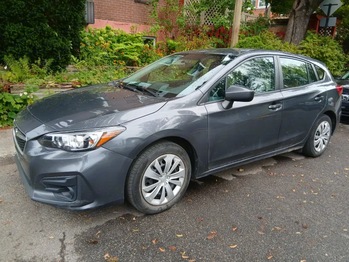 A gray Subaru Impreza hatchback parked on a residential street, not an rv rental. It's an urban scene with green foliage and a brick house.. Subaru Impreza 2018