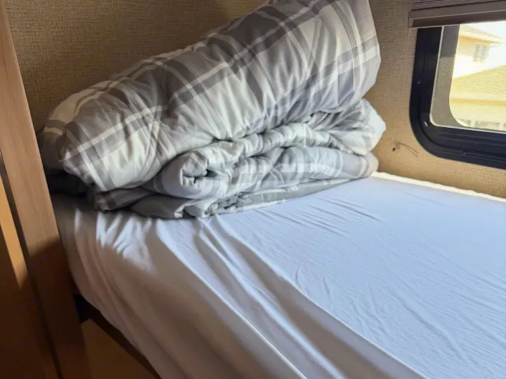 Inside an RV rental: a neatly made bed with a white sheet and rolled gray plaid comforter by the window.. Thor Windsport 2018