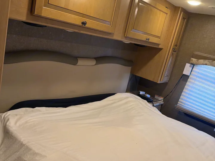 RV rental bedroom: a comfy bed with a headboard, overhead wood cabinets, and light-filtering window blinds.. Thor Windsport 2018