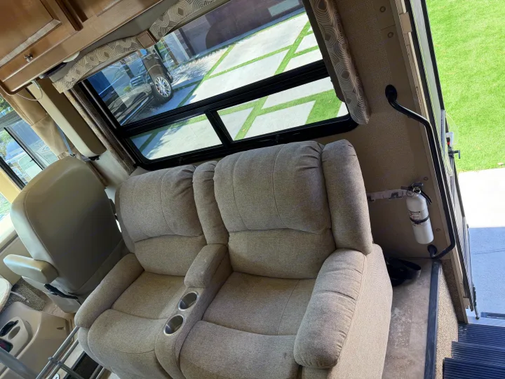 This RV rental features two upholstered recliners with cup holders, a fire extinguisher, and a window view of a grass-patterned driveway.. Thor Windsport 2018