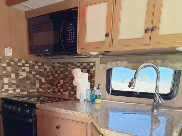 Clean RV rental kitchen featuring a granite-look counter, sink, microwave, stove, and tiled backsplash with cabinets.. Thor Windsport 2018