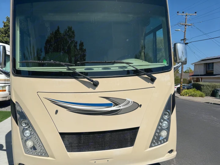 Front view of a cream-colored RV rental, with windshield, wipers, and headlights, parked on a sunny residential street.. Thor Windsport 2018