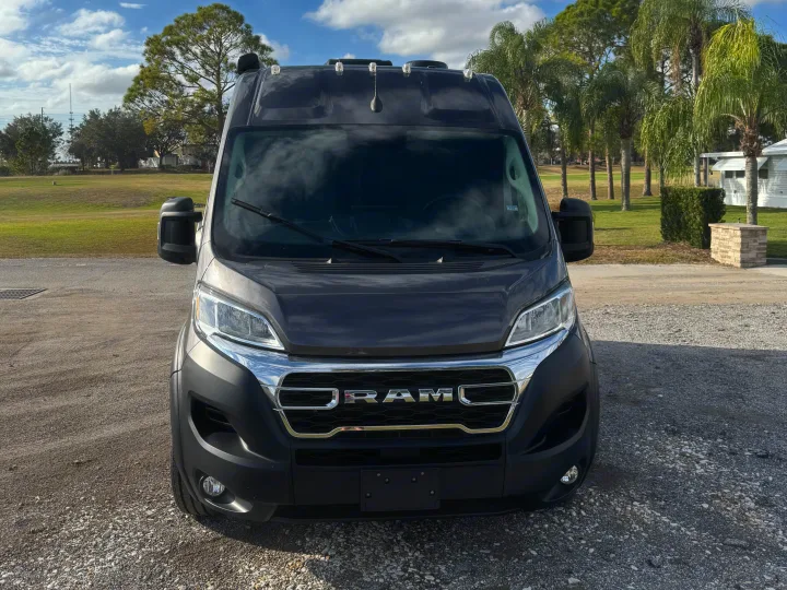 Frontal view: dark gray Ram ProMaster van, an excellent RV rental choice. Parked on gravel with trees and blue sky.. Entegra Coach ARC 2025