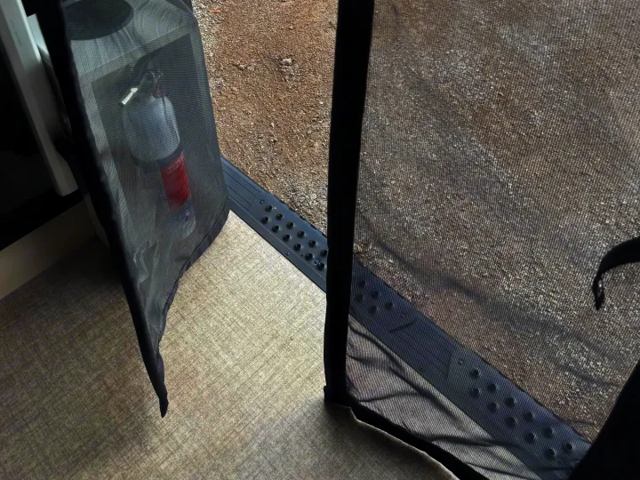 RV rental screen door partially open, showing the gravel outside and a fire extinguisher mounted inside on the interior wall.. Entegra Coach ARC 2025