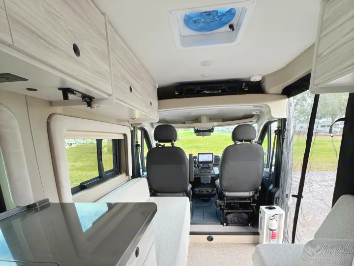 Modern RV rental interior. View from living area to cockpit, showing overhead cabinets, dashboard, seats & roof fan.. Entegra Coach ARC 2025