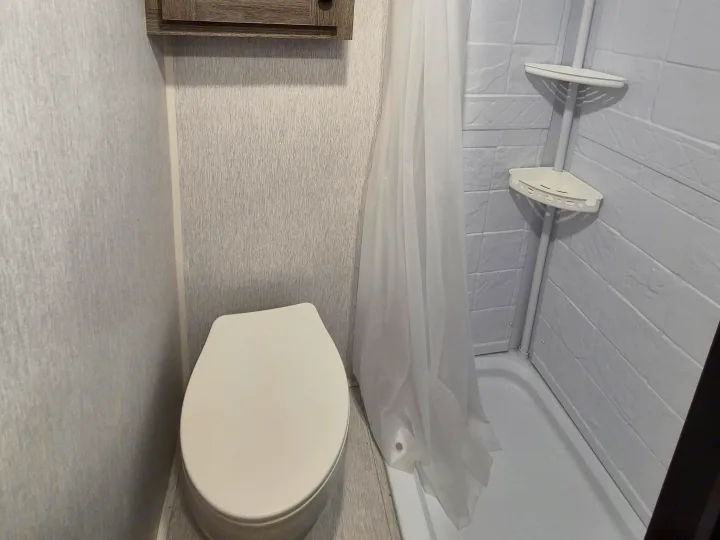 Compact RV rental bathroom featuring a toilet, shower with curtain, and wall-mounted corner shelves for convenience.. Sunlite 16BH 2025