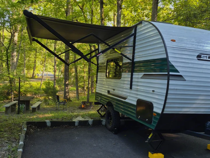An RV rental is set up at a leafy campsite by a stream, complete with a picnic table and a small campfire.. Sunlite 16BH 2025