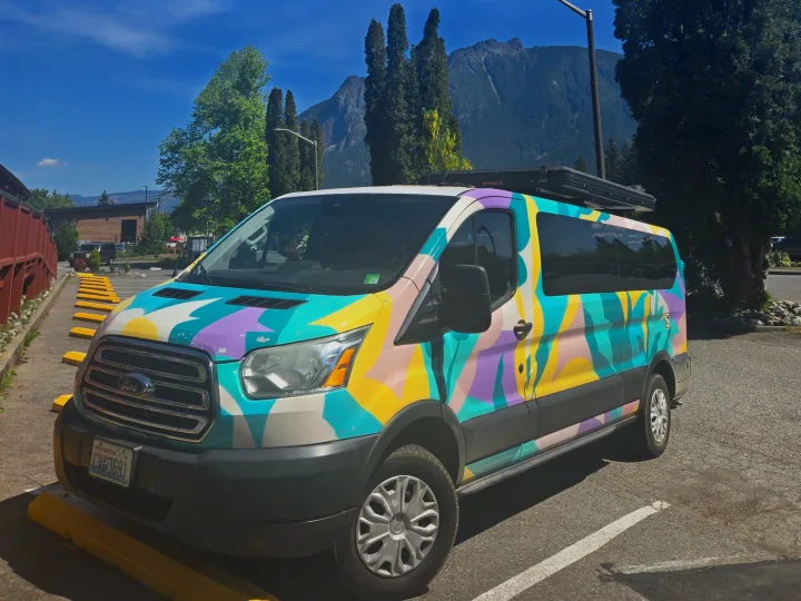 A colorful, abstract-painted Ford Transit RV rental parked against a backdrop of mountains & tall trees under a bright blue sky.. Ford Transit 2017