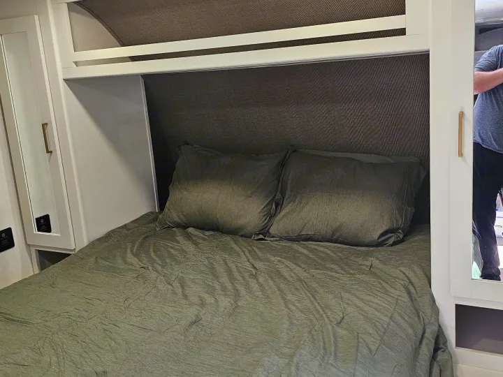 Comfortable RV rental bedroom with a dark green bed, two pillows, and overhead bunk storage, ready for your next trip.. Grand Design Transcend Xplor 2026