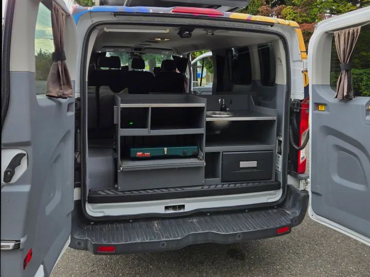 RV rental camper van's custom rear kitchen: sink, portable stove, and storage shelves, with open back doors.. Ford Transit 2017