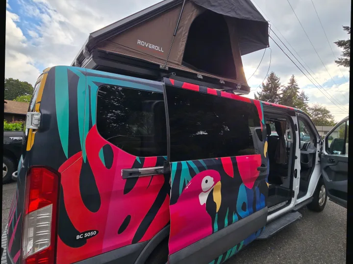 Vibrant RV rental camper van with a tropical parrot wrap, open side door, and a ROVEROLL rooftop tent.. Ford Transit 2017