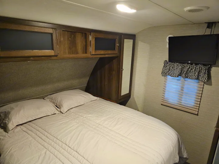 Inside an RV rental, this cozy bedroom features a bed with light bedding, wooden overhead cabinets, a mounted TV, and a window.. Keystone RV Bullet Ultra Lite 2018