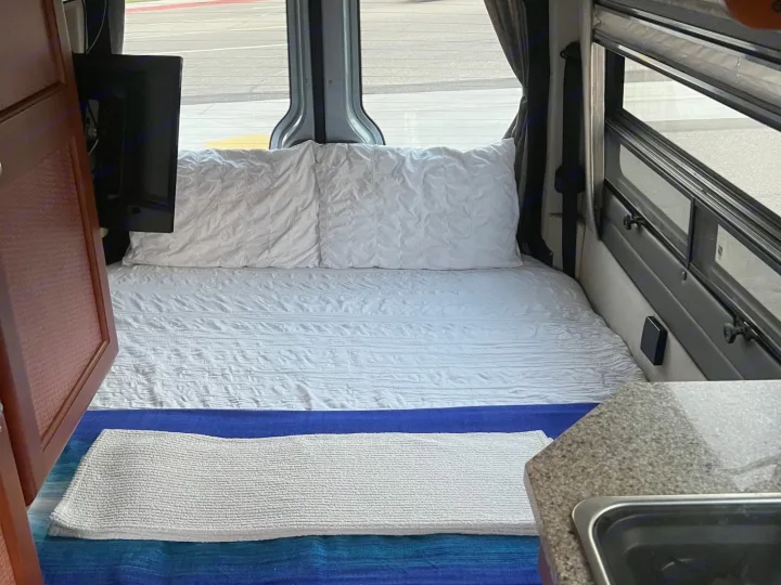 Inside a cozy rv rental: bed with white bedding, wall-mounted TV, and a compact kitchen counter with sink.. Roadtrek Roadtrek Class B 2007