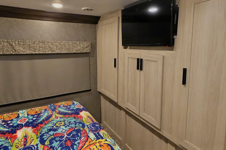 RV rental bedroom with light wood cabinets, TV, and vibrant paisley bedding. A roller blind covers the window.. Forester Classic 2021