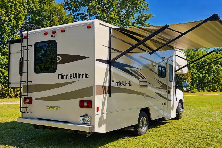 A beige Minnie Winnie RV rental with its awning extended is parked on green grass under a blue sky.. Winnebago Minnie Winnie 2024