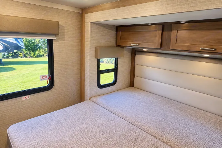 A bright rv rental bedroom featuring a split bed, windows overlooking a green field, and convenient overhead cabinets.. Winnebago Minnie Winnie 2024