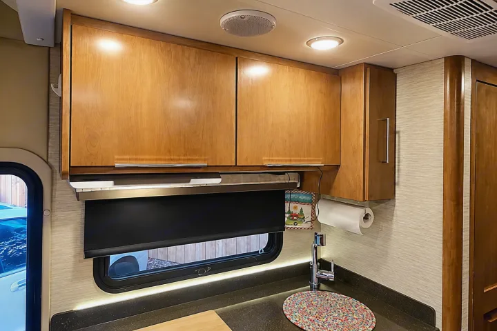 RV rental kitchen: wooden cabinets, sink, dark countertop with under-cabinet lighting, and a window with a blind. Comfortable RV interior.. Tiffin Wayfarer 2020