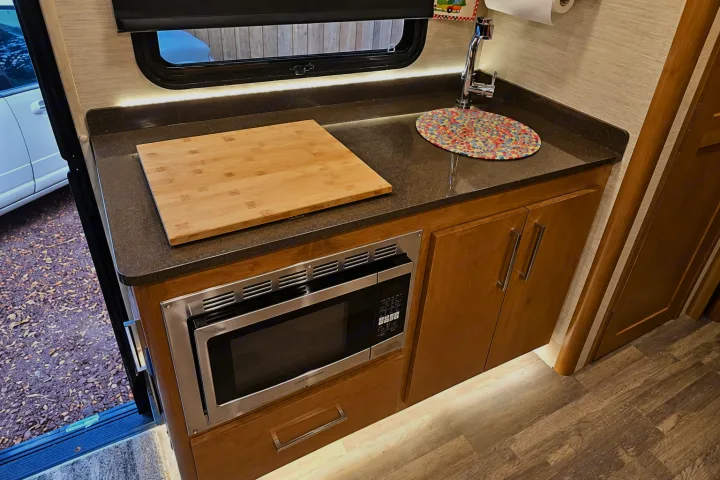 Modern RV rental galley kitchen with under-cabinet lighting, microwave, sink, and wood storage.. Tiffin Wayfarer 2020