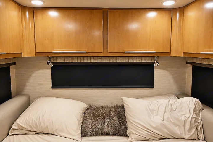 Comfortable RV rental sleeping area featuring wooden overhead cabinets, a window with a black roller blind, and reading lights.. Tiffin Wayfarer 2020