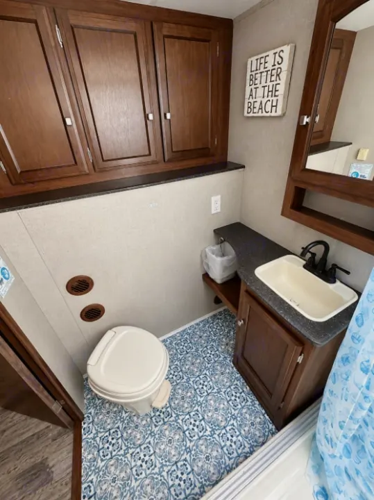 RV rental bathroom with dark wood cabinets, a vanity, toilet, and blue patterned floor. Includes a 'Life is Better at the Beach' sign.. Forest River Freedom 2017